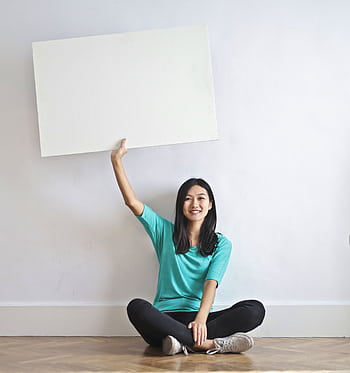 Smiling woman sitting on the floor holding a blank sign indoors, offering copy space. HD phone wallpaper
