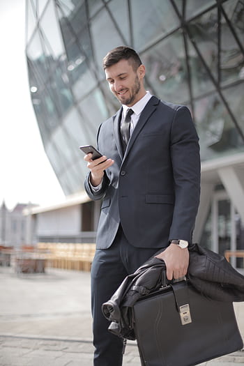 A confident businessman in a suit smiles while texting outside a modern building. HD phone wallpaper