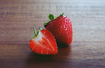 Two fresh strawberries, one whole and one sliced, on a wooden surface. HD wallpaper
