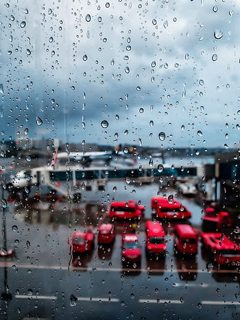 View of an airport through a rain-splattered window, showcasing red vehicles and moody atmosphere. HD phone wallpaper