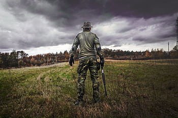 A soldier in camouflage uniform with a rifle stands in a grassy field under a cloudy sky. HD wallpaper
