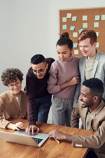 A diverse group of young professionals collaborating around a laptop in a modern office setting. HD phone wallpaper