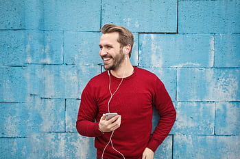 A joyful man in a red sweater smiles while listening to music on his smart against a blue brick wall. HD wallpaper