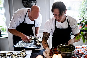 Two chefs in a restaurant kitchen preparing gourmet dishes with fresh ingredients and focused expressions. HD wallpaper