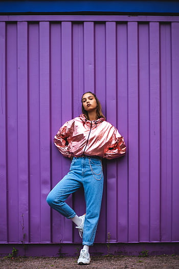 Stylish woman in metallic jacket and jeans posing confidently against a bold purple wall. HD phone wallpaper