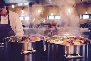 A chef in a restaurant kitchen tends to large pots of steaming soup, creating a cozy atmosphere. HD wallpaper