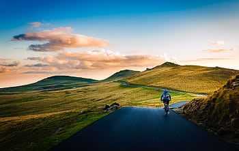 Cyclist biking on a scenic rural road with rolling hills at sunset. HD wallpaper