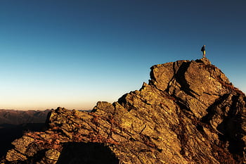 A lone hiker stands triumphantly atop a rocky mountain peak at sunrise, embracing nature and the view. HD wallpaper