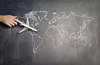 A hand guides a toy airplane over a world map drawn in chalk on a blackboard, symbolizing travel planning. HD wallpaper