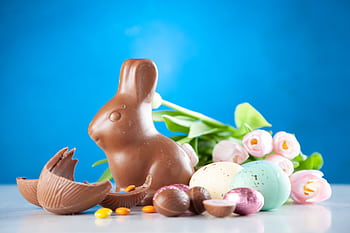 A delightful Easter scene with a chocolate bunny, colorful eggs, and pink tulips on a blue background. HD wallpaper
