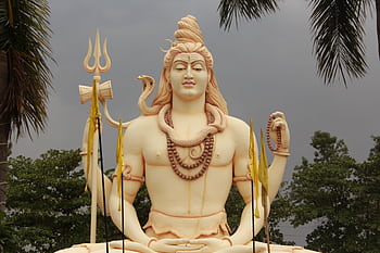 A grand Shiva statue amidst palm trees with a dramatic cloudy backdrop. HD wallpaper