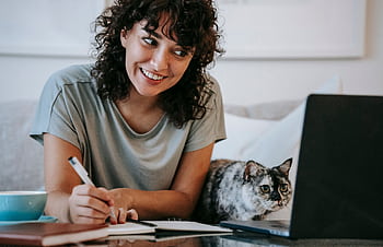 Happy woman writing notes near laptop with cat at home, enjoying lance work environment. HD wallpaper
