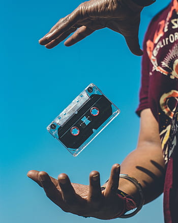 A retro cassette tape floating between hands against a clear blue sky, evoking nostalgia. HD phone wallpaper