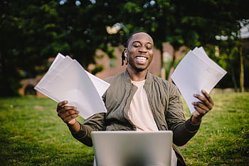 Happy young man holding papers with a laptop outdoors, showcasing academic achievement and joy. HD wallpaper