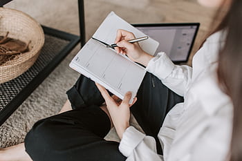 A young woman sitting on the floor at home, writing in a planner with a pen, showcasing a casual lifestyle. HD wallpaper