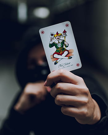 Mysterious hooded figure holding a joker card in a dimly lit setting, emphasizing intrigue and drama. HD phone wallpaper