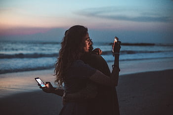 Couple embracing on a beach at sunset, using mobile s, symbolizing modern love. HD wallpaper
