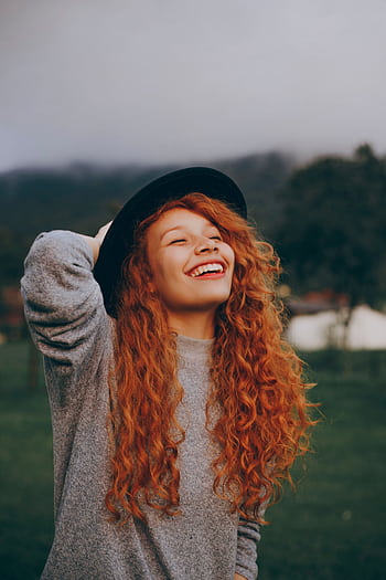 A happy young woman with curly red hair and a hat smiling in a lush green park. HD phone wallpaper