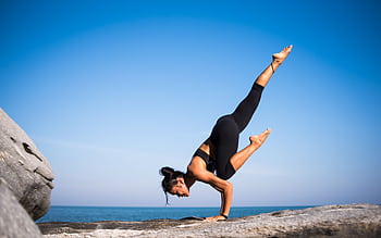 Woman performing a yoga pose on a rocky beach with ocean view and clear blue sky. HD wallpaper