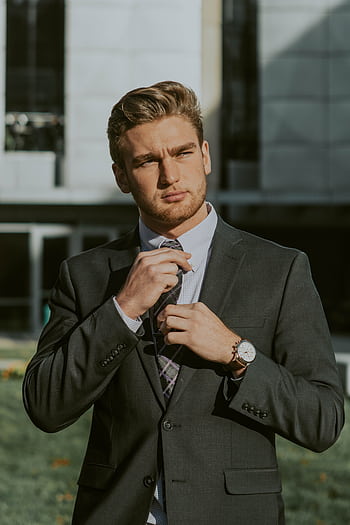A young businessman in a suit confidently adjusts his tie outdoors with a modern building in the background. HD phone wallpaper