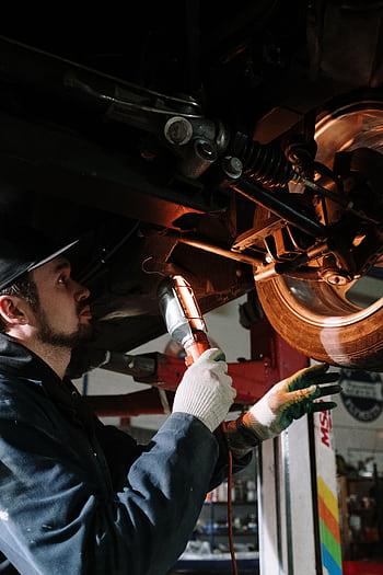 Mechanic uses a flashlight to inspect car's suspension in a garage setting. HD phone wallpaper