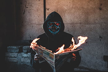 A hooded figure with a glowing mask holds a burning newspaper inside a dim room. HD wallpaper