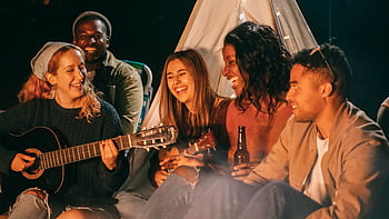 A diverse group of friends playing guitar and ukulele by a campfire at night, having fun and socializing. HD wallpaper
