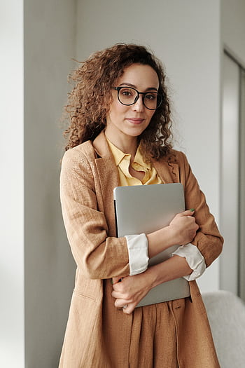 A confident woman in formal attire holding a laptop, standing in an office environment and looking at the camera. HD phone wallpaper