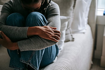 A woman sits on a bed, hugging her knees in a contemplative pose, indoors. HD wallpaper