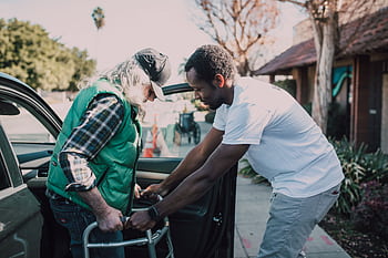 Young volunteer helping senior man with walker near a parked car, symbolizing care and support. HD wallpaper