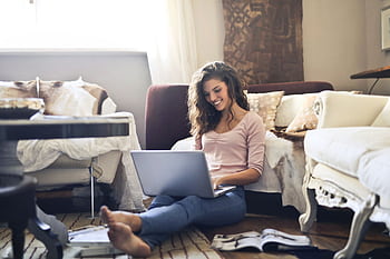 Smiling woman using a laptop seated on the floor in a cozy living room, working remotely. HD wallpaper