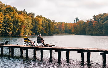 Two men fishing on a pier surrounded by autumn foliage at a scenic lake. HD wallpaper