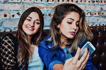 Two young women capturing a selfie while sitting in a casual indoor setting, smiling and relaxed. HD wallpaper