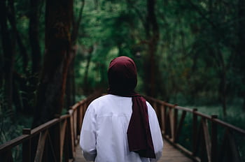 A woman in a hijab walks on a wooden bridge surrounded by lush greenery in a tranquil forest setting. HD wallpaper