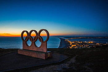 Scenic sunset view of the Olympic rings monument by the sea with city lights in the distance. HD wallpaper