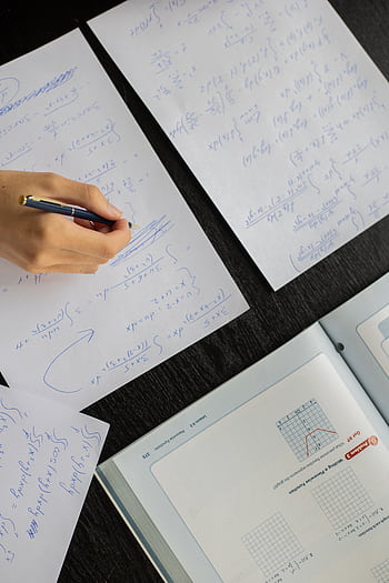 Close-up of a student solving mathematical equations with papers and textbook on a table. HD phone wallpaper