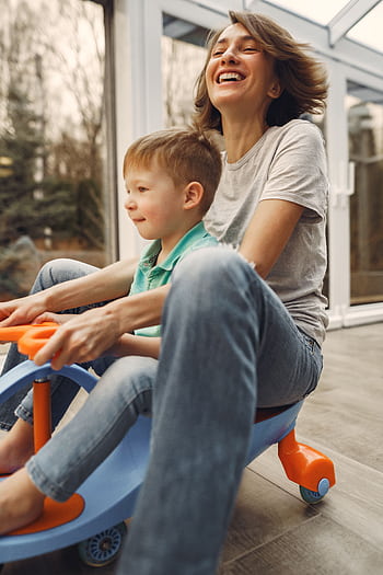A joyful mother and son playing together on a toy car inside a sunlit room. HD phone wallpaper