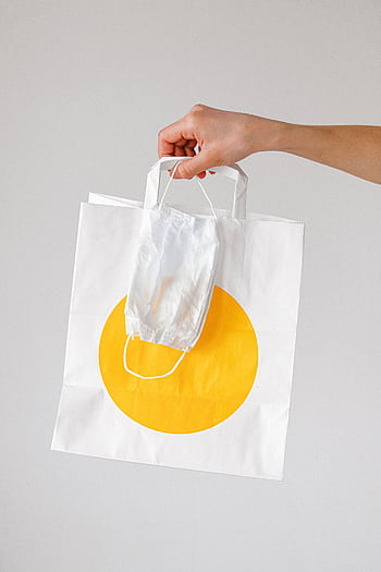Close-up of a hand holding a paper shopping bag with a face mask, symbolizing pandemic shopping habits. HD phone wallpaper