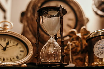 Close-up of a vintage hourglass surrounded by antique clocks in warm lighting. HD wallpaper