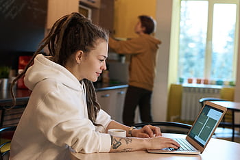 Young woman with dreadlocks works on a laptop in a casual kitchen environment. HD wallpaper