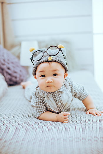 Adorable baby wearing a knit beanie and glasses on a bed, showcasing innocence and charm. HD phone wallpaper