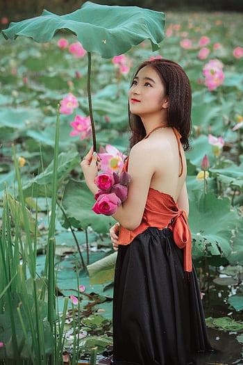 A stylish Asian woman stands amidst blooming lotus flowers, holding a bouquet and a large leaf. HD phone wallpaper