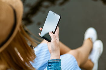 A young woman holding a smart while sitting outdoors in summer, capturing a moment of relaxation and technology use. HD wallpaper