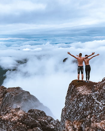 Two hikers stand on a rocky cliff, embracing the expansive mountain view and clouds, symbolizing dom and exploration. HD phone wallpaper