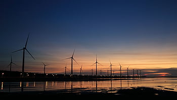Serene view of wind turbines reflecting on water at sunset, symbolizing renewable energy. HD wallpaper