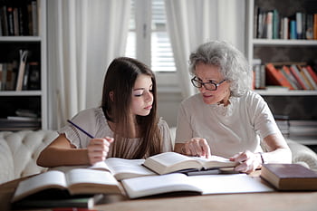 A grandmother helps her teenage granddaughter with homework, surrounded by books indoors. HD wallpaper