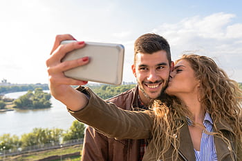 A happy couple taking a romantic selfie by the scenic riverside on a sunny day. HD wallpaper