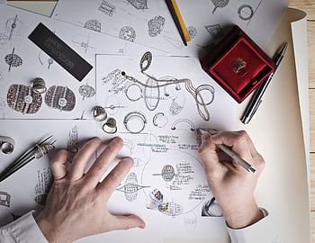 Hands sketching jewelry designs with tools and prototypes on a desk. HD wallpaper