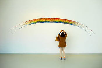 A woman stands observing a colorful rainbow art installation indoors. HD wallpaper