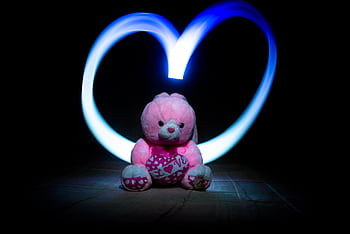 Adorable pink teddy bear with heart-shaped light painting in a dark setting, symbolizing love. HD wallpaper